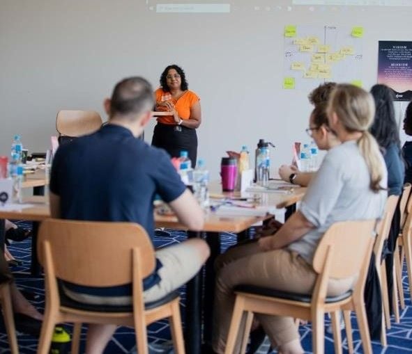 Renée Madsen facilitating a team reconnection session, standing in front of a group of seated team members in a conference room.
